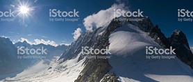 Dramatic sunlight flaring over snow capped Alpine mountain peaks, glaciers and rocky pinnacles as mountaineers, dwarfed by the scale of the environment, steadily ascend the cloud shrouded summit of Aiguille Verte in the Mont Blanc massif, Haute-Savoie, France. ProPhoto RGB profile for maximum color fidelity and gamut.