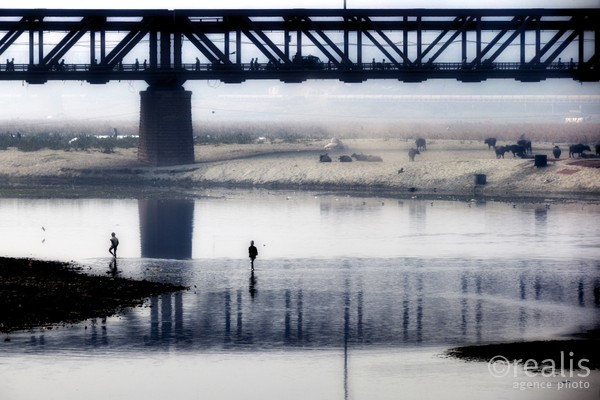 India Follows Raabe 08 - Zwei Männer in einem Fluss in Indien, der von einer Brücke überquert wird, auf der Menschen unterwegs sind
