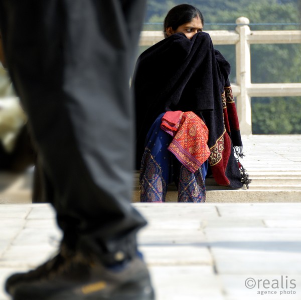 India Follows Raabe 20 - Eine indische Frau versteckt mit ihrem Schleier ihr Gesicht vor einem vorbeikommenden Mann, dessen Beine im Vordergrund zu sehen sind.