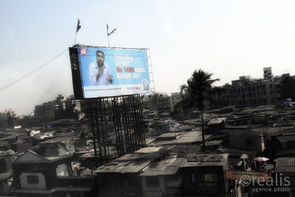 India Follows Raabe 22 - Blick auf die Slums von Bombay über denen ein großes Werbeplakat schwebt und zum Konsum animiert.
