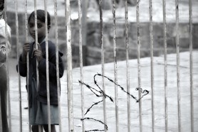 India Follows Raabe 23 - Ein Kind aus Indien steht verloren und zugleich fragend hinter einem Gitter. Ein Stück Stacheldraht dokumentiert zusätzlich seine Hofnungslosigkeit.