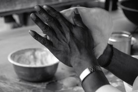 India Follows Raabe 28 - Ein indischer Bäcker bei seiner Arbeit. Eine Nahaufnahme seiner feingegliederten, geschmeidigen und dunklen Hände, zwischen denen der Fladen entlanggleitet. Eine Schale mit Mehl steht in der Nähe.