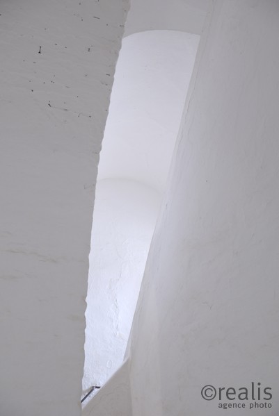 Hold - Blick in ein weißes Treppenhaus, in dem - eingerahmt von den weißen Wänden - das Endstück eines Handlaufs zu sehen ist. Grafische Wirkung