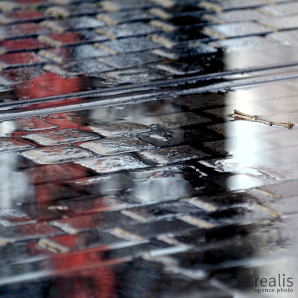 Black meets Red - Ein roter und ein schwarzer Schatten von Menschen spiegeln sich auf dem regennassen Pflaster einer Straße, durch die die Schienen einer Straßenbahn verläuft.
