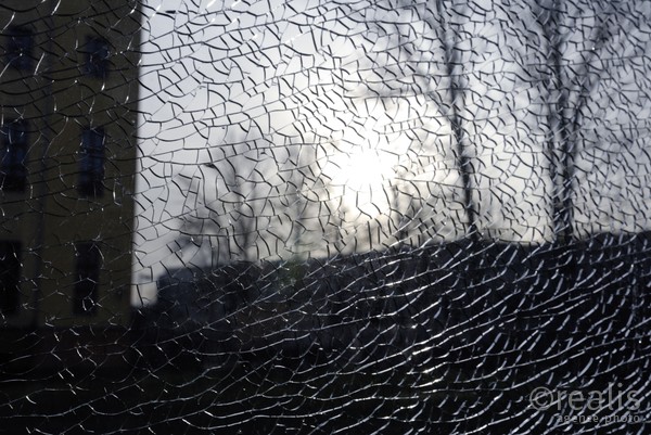 Fragile world - Hinter der zerbrochenen Glasscheibe zeigt sich die fragile Struktur einer Landschaft mit Sonne, Bäumen und Häuserfassade.
