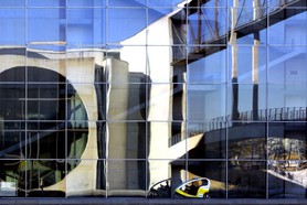 Outsider - In einem Regierungsgebäude in Berlin mit seinen riesigen Glasfassaden und einem Einblick ins Innere des Gebäudes spiegeln sich die gegenüberliegenden Fassaden, Passanten und ein vorbeifahrendes Fahrrad-Taxi.