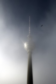 Raising - Der Alexanderturm in Berlin wird vom Nebel umgeben, der von der Sonne verdrängt und von einem Vogel beobachtet wird.
