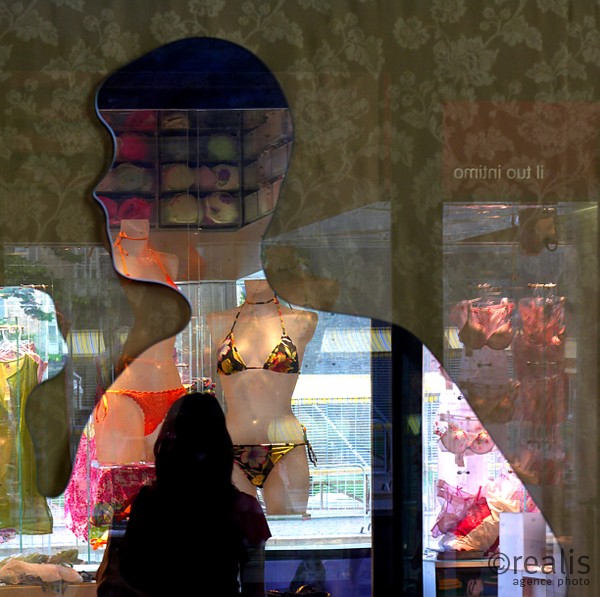 Travel sick - Eine Frau steht vor einem Schaufenster mit ausgestellten Bikinis und dem Schattenbild aus Pappe eines Mannes. Koffer packen und verreisen könnte dem Betrachter in den Sinn kommen