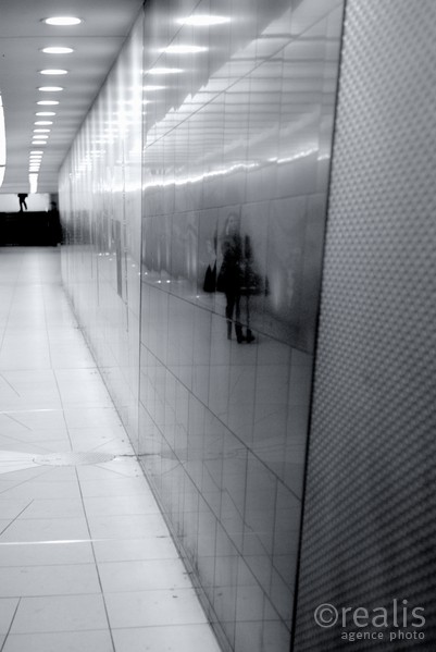 Underground - Das Spiegelbild einer telefonierenden Frau wird in einem U-Bahngang von einem Mann verfolgt, dessen Beine am oberen Bildrand zu sehen sind.