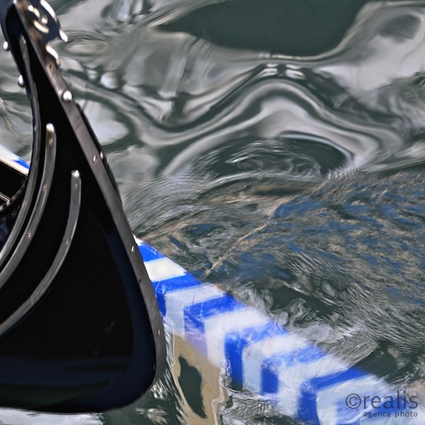 Venice charm - Die schwarze Vorderseite einer venezianischen Gondel spiegelt sich mit dem Vorderteil eines blau-weißen Paddels im Wasser und erzeugt eine sprudelnde, lebendige Gestalt des sie umgebenden Wassers.